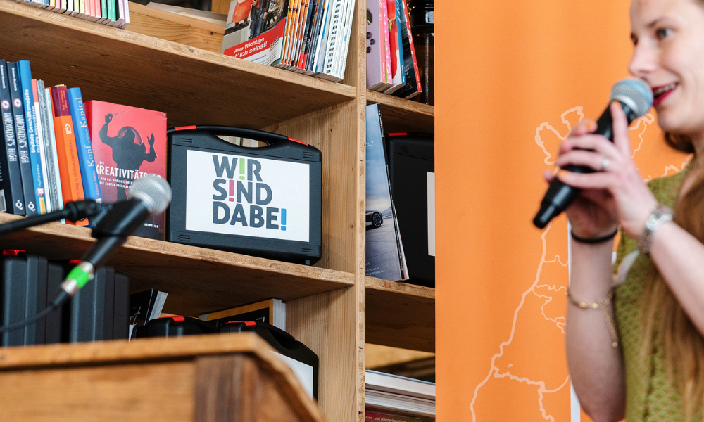 Eine Frau spricht in ein Mikrofon. Im Hintergrund steht ein WSD-Koffer im Bücherregal.