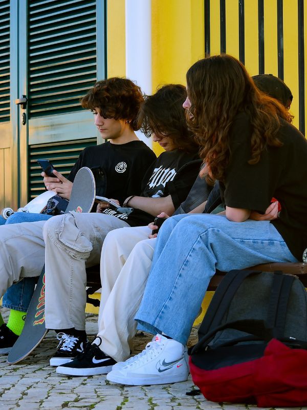 Eine Gruppe von zwei Jungen und zwei Mädchen sitzt auf einer Bank, sie haben Skateboards dabei. Zwei der Jungs schauen etwas auf ihrem Handy nach.
