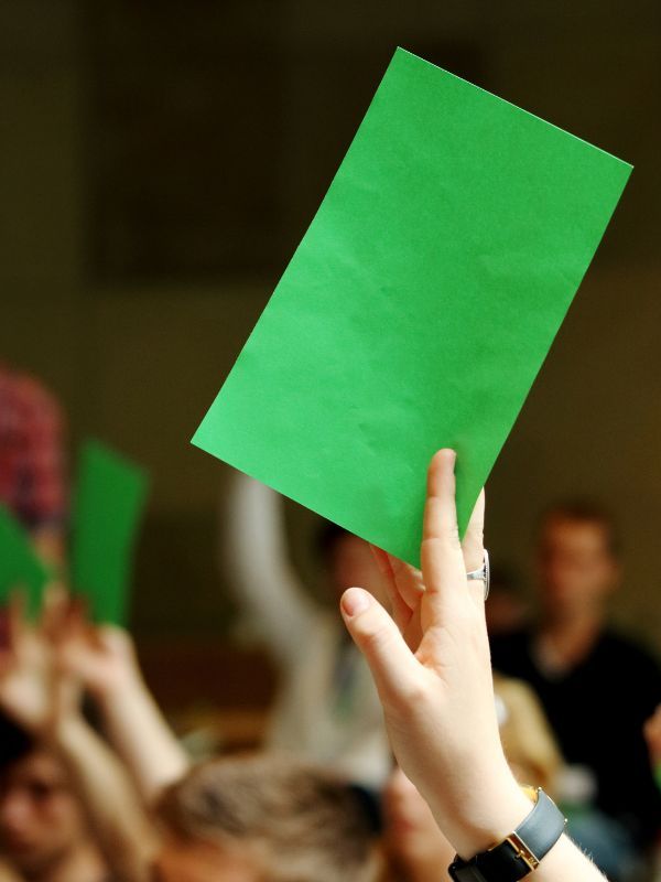 Der Hintergrund zeigt verschwommen eine Veranstaltung mit vielen Menschen. Im Vordergrund hebt eine junge Frau eine grüne Karte in die Luft.