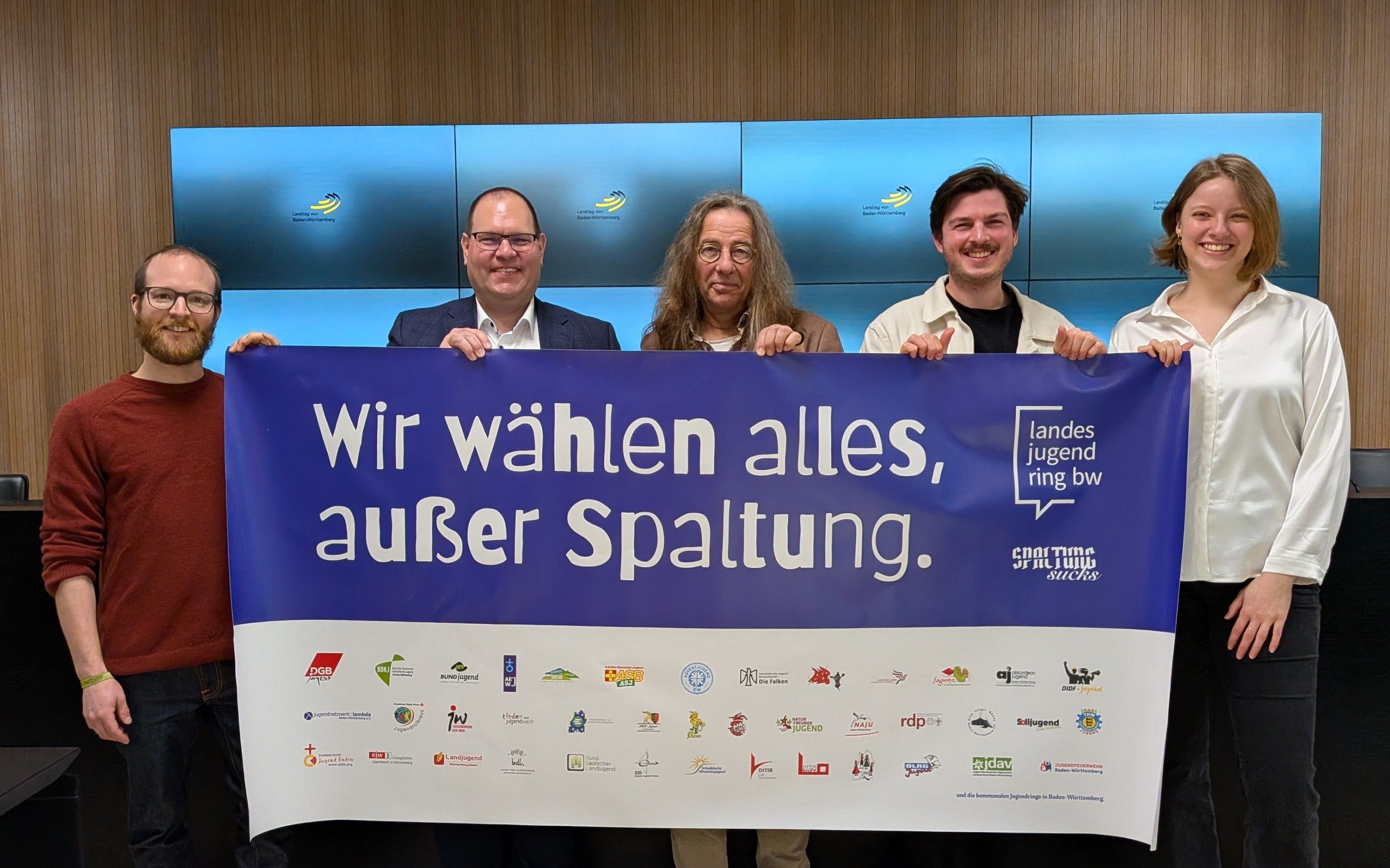 Auf dem Bild sind vier Männer und eine Frau zu sehen, die ein Transparent halten. Darauf steht: Wir wählen alles, außer Spaltung.