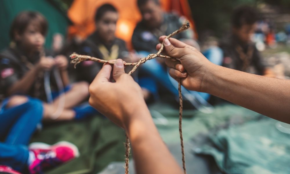 Im Vordergrund ist ein junger Mensch zu sehen, der vorführt wie man einen Knoten schlingt. Im Hintergrund sind Kinder, die den Knoten nachmachen.