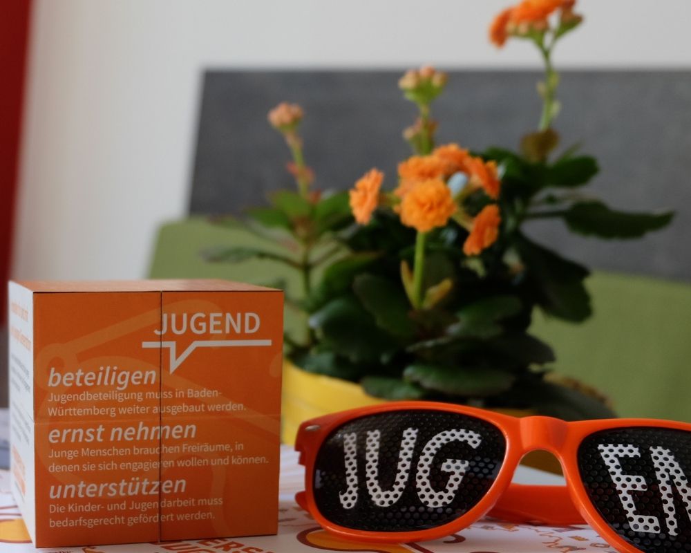Auf einem Tisch steht eine Blume mit orangefarbenen Blüten. Davor liegt eine Brille, auf deren Gläsern das Wort Jugend abgebildet ist, daneben liegt ein Würfel, auf dem als Motiv ein Text steht: Jugend beteiligen, ernst nehmen, unterstützen.